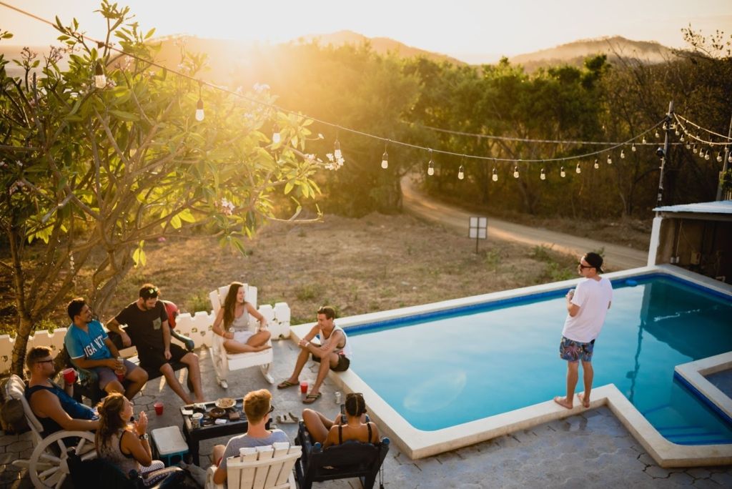 Eight people sat in a circle eating and drinking next to a swimming pool, another man is slightly away from the group, standing on the edge of the pool looking at the group.