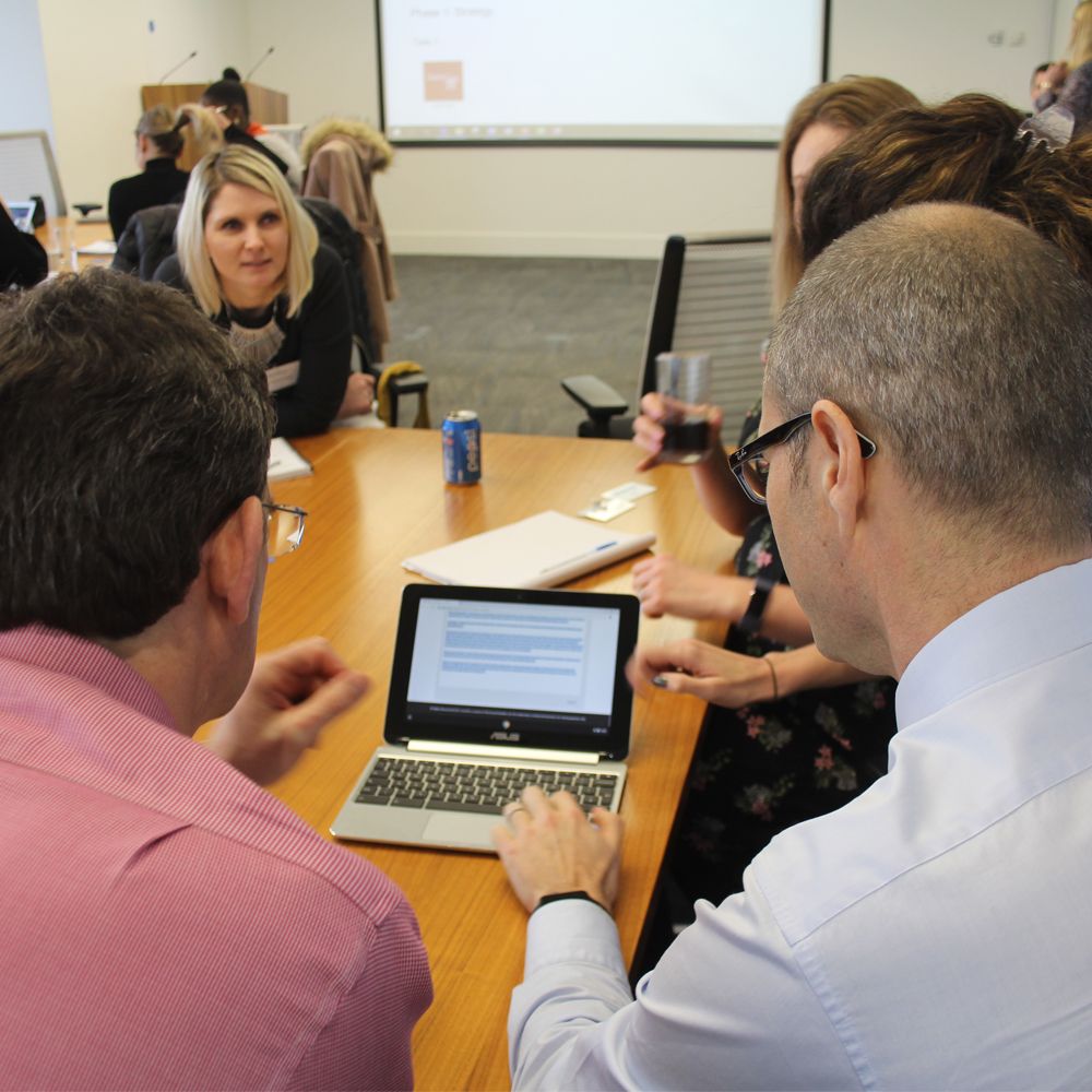 Team of people sat around table looking at laptop, Crisis90 training platform on the screen